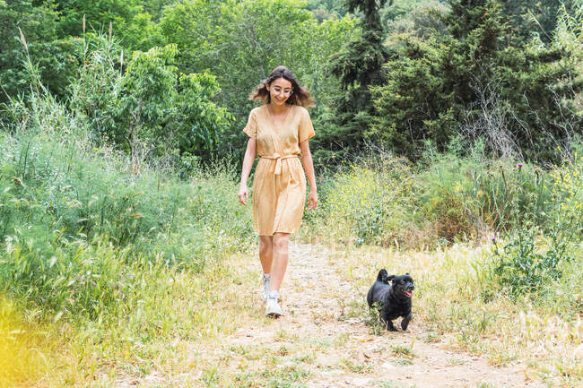 Счастливая женщина в летнем платье гуляет с черной симпатичной собакой в парке и наслаждается выходными вместе — стоковое фото
