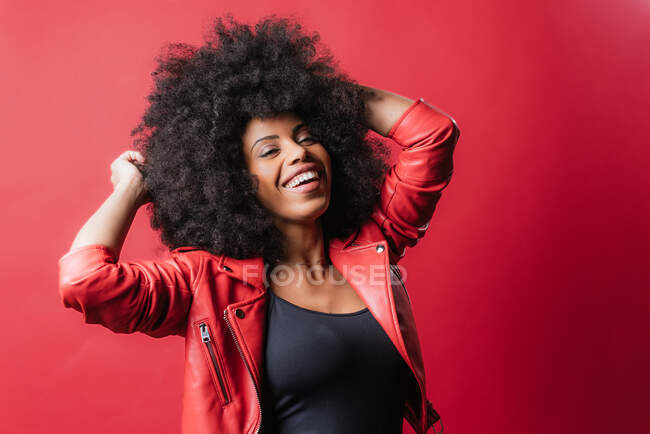 Carefree African American female with Afro hairstyle touching hair with closed eyes on red background in studio — Stock Photo