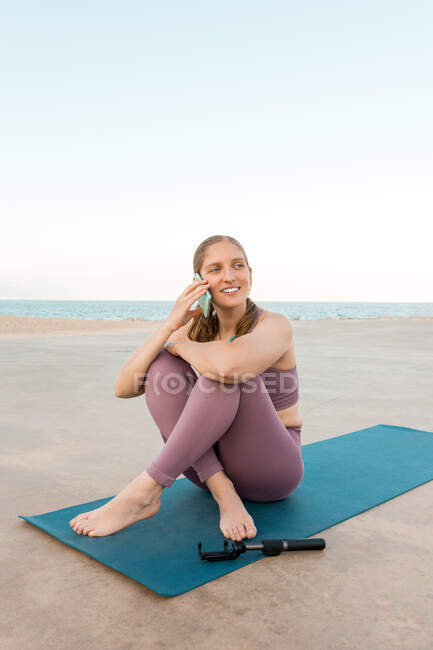 Mulher encantada em activewear sentado no tapete de ioga e falando no telefone celular na praia enquanto olha para longe — Fotografia de Stock