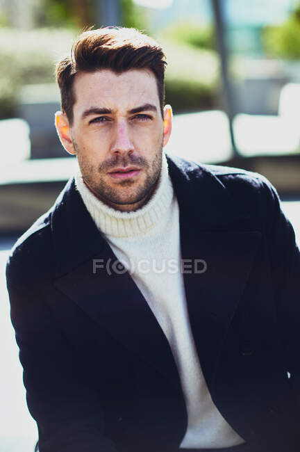 Adulto maschio barbuto in maglione e cappotto per strada guardando la fotocamera in città durante il giorno — Foto stock