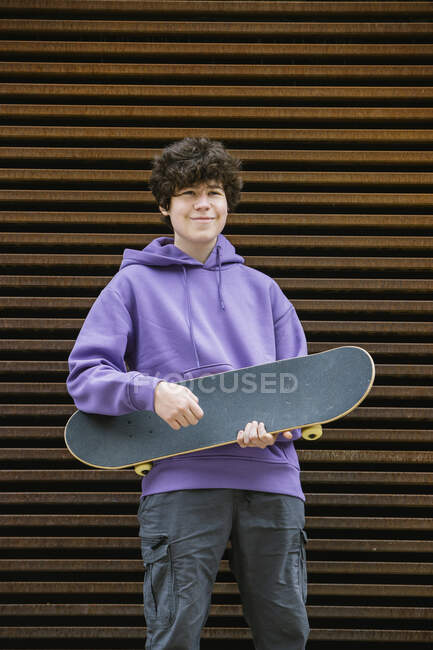 Щасливий молодий чоловік в повсякденному одязі зі скейтбордом, дивлячись далеко, стоячи біля стіни на вулиці — стокове фото