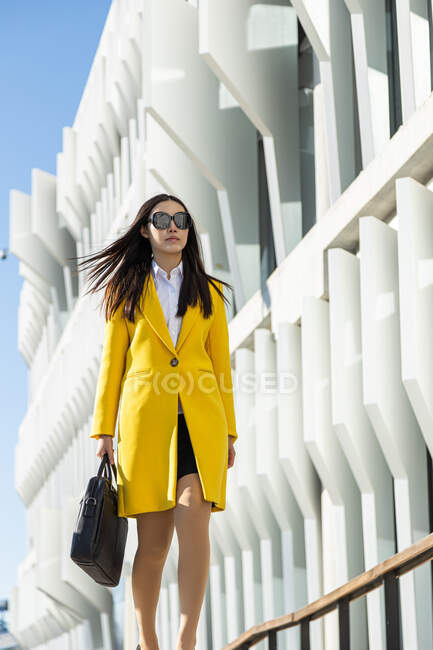 Asiatische Geschäftsfrau mit gelbem Mantel und Smartphone läuft auf der Straße, im Hintergrund ein Gebäude — Stockfoto
