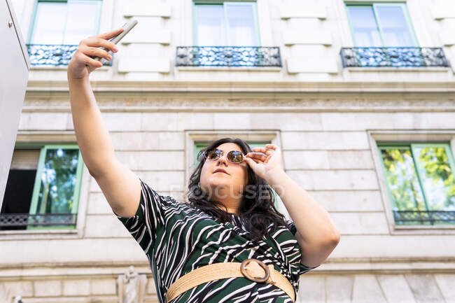 Снизу молодая женщина плюс размер в стильном платье и солнцезащитные очки делая селфи на смартфоне, стоя рядом с каменным зданием в городе — стоковое фото