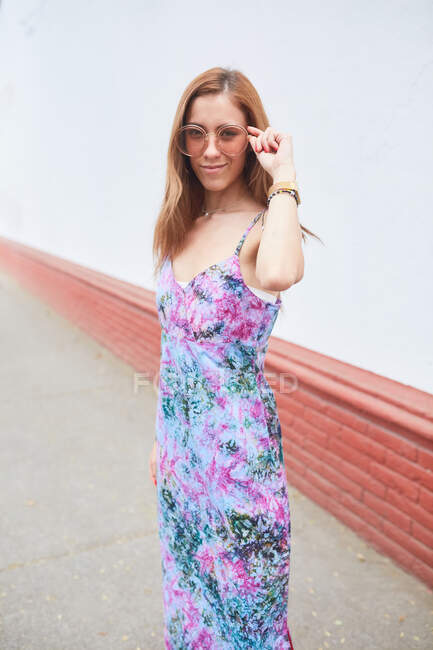 Весела жінка в модних сонцезахисних окулярах і літній сукні стоїть на вулиці і дивиться на камеру — стокове фото