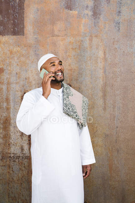 Веселый мусульманский мужчина в традиционной одежде улыбается и просматривает мобильный телефон, стоя рядом с потрепанной стеной на улице — стоковое фото