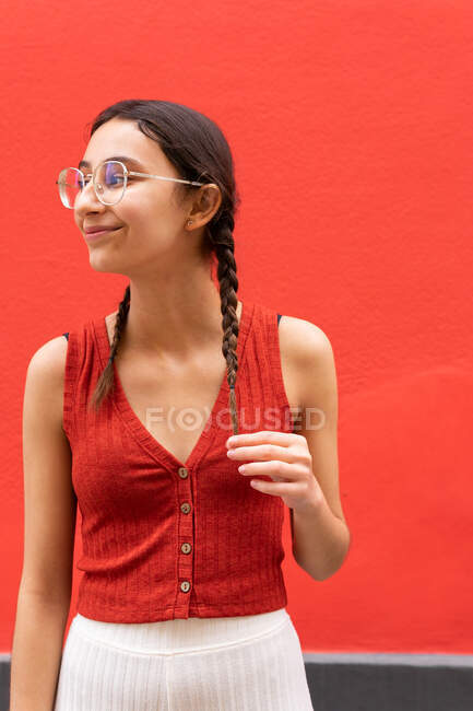 Fröhliche junge Frau berührt Zöpfe, während sie auf rotem Hintergrund in der Straße wegschaut — Stockfoto