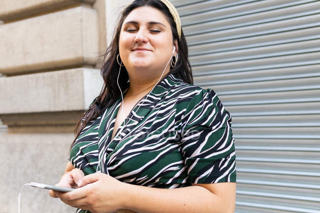 Glückliche junge kurvige Frau in stylischem Zierkleid und Stirnband hört Audio-Nachrichten per Handy mit Kopfhörer und lacht, während sie an der Wand eines modernen Gebäudes steht — Stockfoto