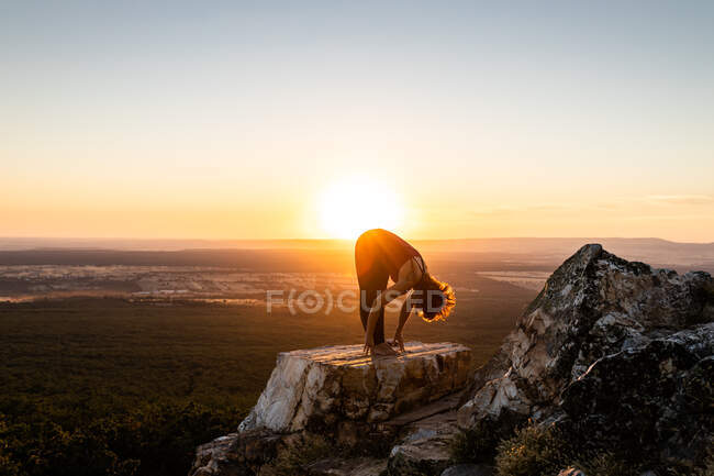 Joven yogi practicando yoga sobre una roca en la montaña con la luz del amanecer, vista lateral sosteniendo los tobillos - foto de stock
