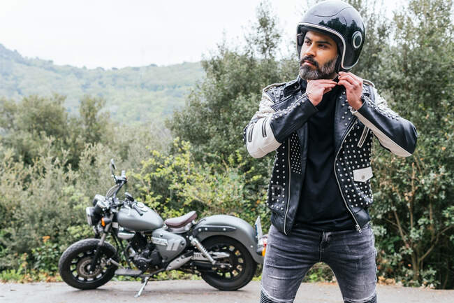 Self assured adult bearded male biker in stylish leather jacket adjusting protective helmet and looking away in nature — Stock Photo