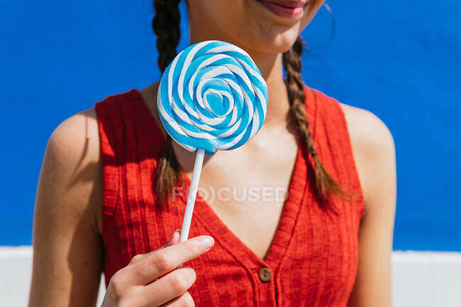 Crop of anonymous female standing with sweet lollipop in street on sunny day on blue background and looking away — Stock Photo
