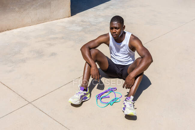 High angle of tired African American male athlete sitting with jumping rope on sports ground and taking break during workout in summer — Stock Photo