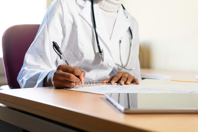 Ernte unkenntlich schwarze Ärztin mit Stethoskop, die Informationen auf Papierbogen schreibt, während sie medizinischen Bericht am Tisch im Büro der modernen Klinik vorbereitet — Stockfoto