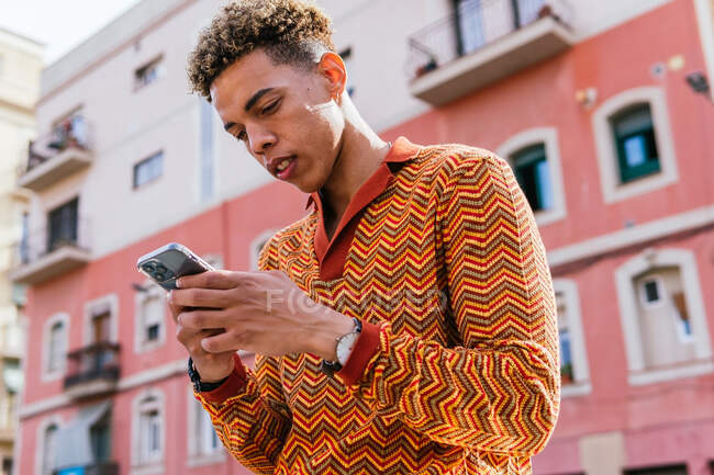 Von unten Seitenansicht eines jungen hispanischen Typen mit Afro-Haaren in stylischem, buntem Outfit, der im Sonnenlicht auf einem Geländer in der Nähe eines städtischen Gebäudes steht und sein Handy surft — Stockfoto