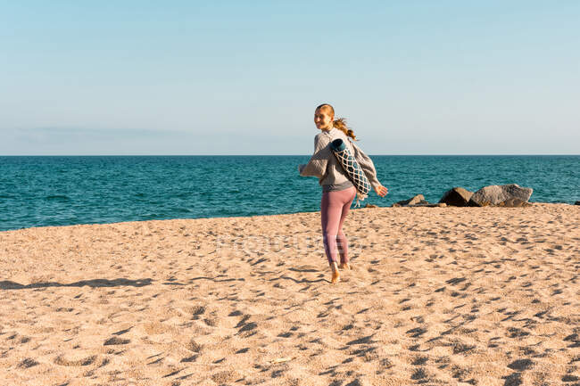 Full body side view of young happy barefoot female in activewear with rolled yoga mat looking at distance over the shoulder to the camera while standing on sandy beach near sea — Photo de stock