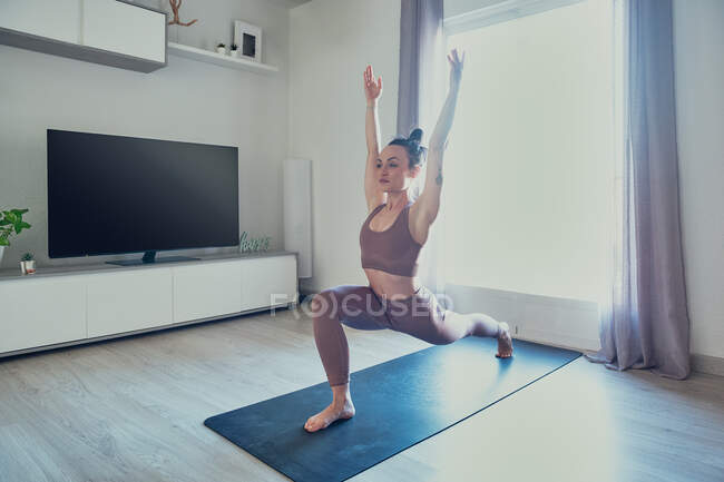 Молода гнучка жінка в спортивному одязі, що практикує йогу з піднятими руками, дивлячись вперед в домашній кімнаті на задньому світлі — стокове фото