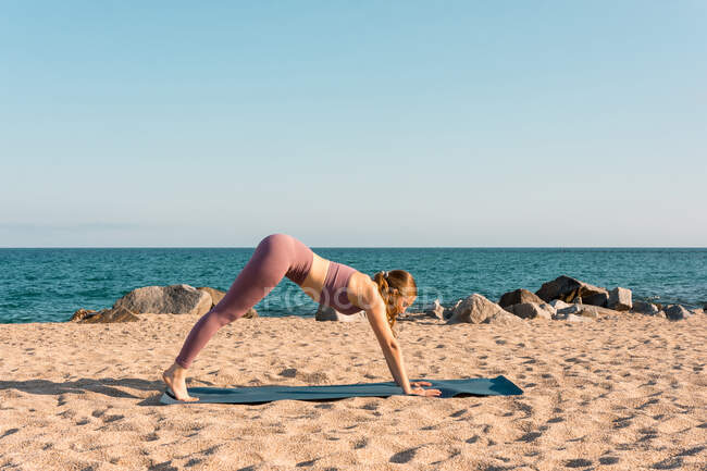 Vista laterale di flessibile femminile praticare yoga in Adho Mukha Svanasana sulla spiaggia sabbiosa vicino al mare — Foto stock