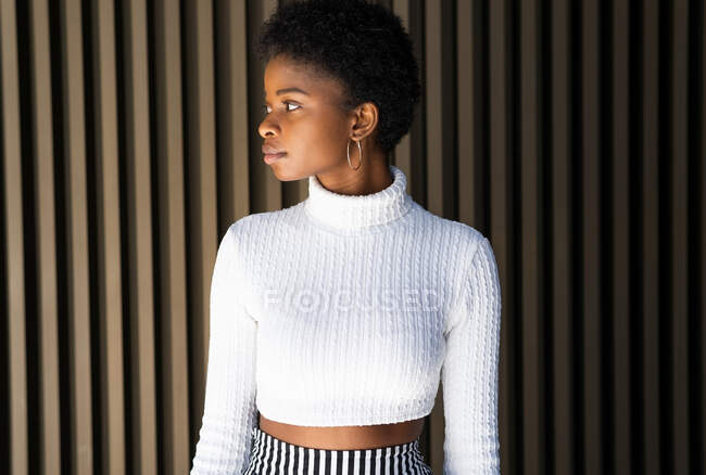 Mujer afroamericana sin emociones en suéter de moda mirando hacia otro lado contra la pared del edificio a rayas en la calle - foto de stock