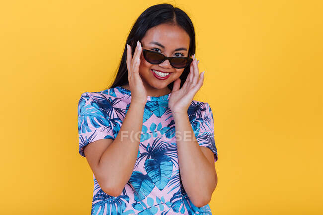 Glückliche asiatische Frau in stylischer Sonnenbrille und T-Shirt mit tropischem Blatt-Print schaut in die Kamera auf gelbem Hintergrund im Studio — Stockfoto