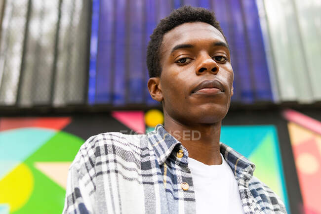 Dal basso grave sognante maschio afro-americano in piedi guardando la fotocamera sfondo colorato in strada — Foto stock