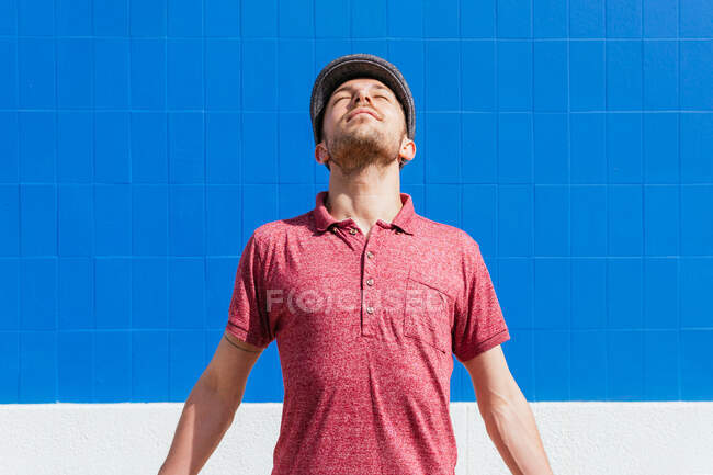 Heureux jeune homme barbu en polo rouge et chapeau debout avec les yeux fermés contre le mur bleu et profiter de la liberté et de la lumière du soleil dans la journée d'été — Photo de stock