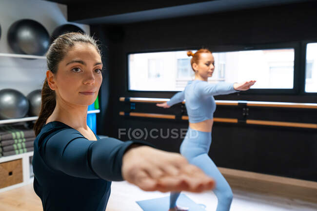 Focused young female instructor standing in Warrior II position while practicing yoga with student in modern fitness studio — Stock Photo