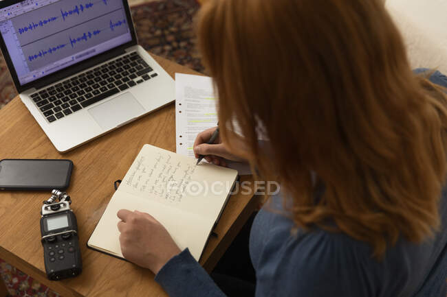 Зверху анонімної жінки роблять нотатки в блокноті, сидячи за столом з мікрофоном і ноутбуком і готуючись до запису подкасту — стокове фото