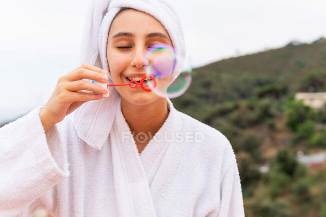 Junge Frau in weißem Bademantel und Handtuch bläst Seifenblasen bei Wellness-Session auf Balkon — Stockfoto