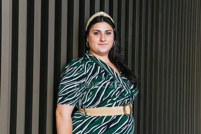 Молодая кривая женщина модель в стильном черном платье с диагональю полосатый отпечаток и пояс, стоящий против стеклянной стены на улице — стоковое фото