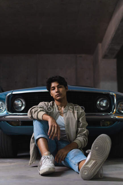 Портрет молодого латинця в повсякденному одязі, впевнено дивлячись на камеру, сидячи спираючись на старовинну машину на парковці — стокове фото