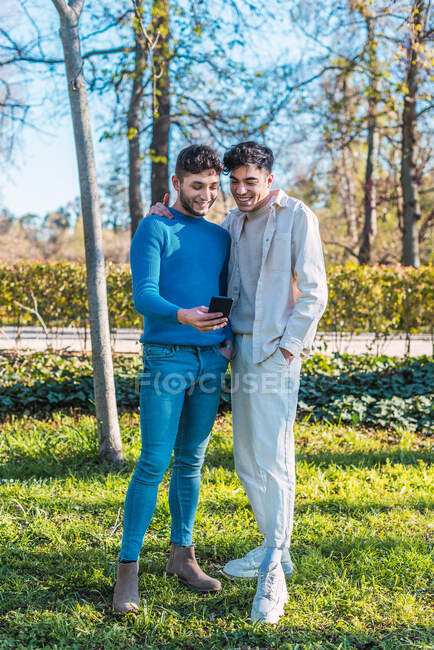 Delighted homosexual couple of males embracing and watching funny video on mobile phone while standing in park and having fun — Stock Photo