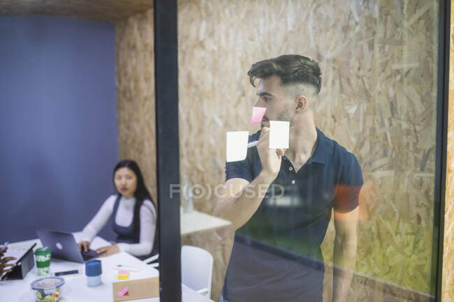 Administrador executivo masculino escrevendo em nota pegajosa na parede de vidro durante brainstorm com colegas de trabalho no escritório — Fotografia de Stock