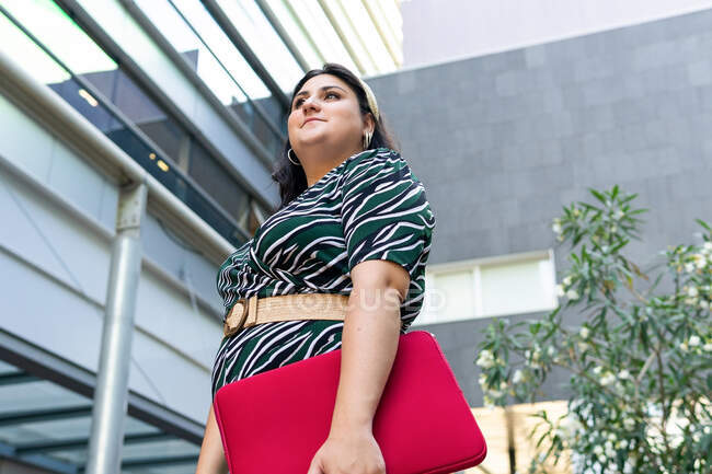 Side view of cheerful young female in stylish striped outfit with red laptop case in hands smiling while standing against contemporary city building — Stock Photo
