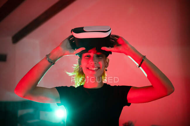 Mulher feliz vestindo t-shirt ao usar óculos VR e de pé olhando para a câmera em estúdio com luzes de néon vermelho — Fotografia de Stock