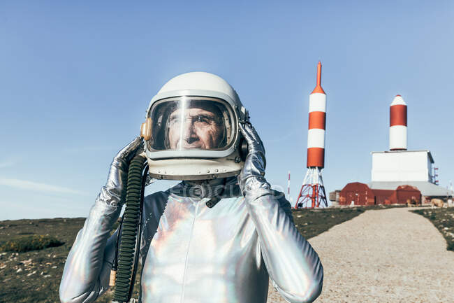 Senior man in spacesuit taking off protective helmet while standing on path near spaceport with rocket shaped antennas on cloudless day — Stock Photo