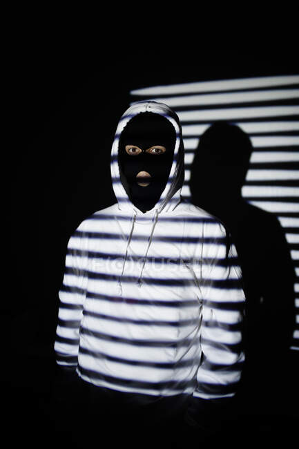 Unrecognizable serious rebellious young male in black balaclava mask and white hoodie standing near wall with jalousie shadow — Stock Photo