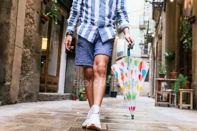 Crop unrecognizable male in summer outfit walking with umbrella on wet street in city after rain — Stock Photo