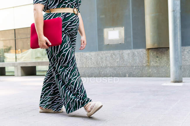 Side view of anonymous cheerful young female in stylish striped outfit with red laptop case in hands while walking against contemporary city building — Stock Photo