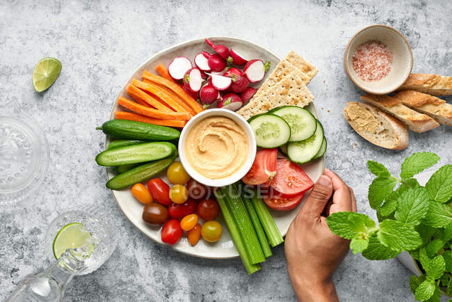 From above crop person holding of appetizing fresh raw cut cucumbers with carrots and radish served on white table with assorted tomato and hummus and placed on green table with pieces of bread — Stock Photo
