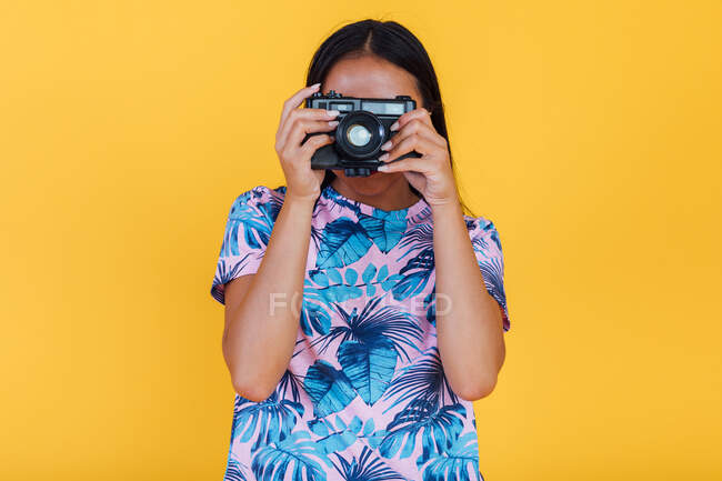 Анонимная женщина в футболке с тропическим отпечатком листьев, фотографирующая на фотокамеру на жёлтом фоне в студии — стоковое фото