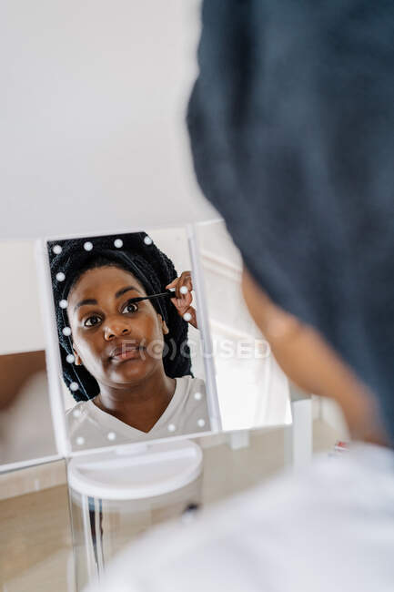 Reflejo del espejo de la hembra afroamericana adulta aplicando rímel cosmético en las pestañas durante el procedimiento de maquillaje diario en casa - foto de stock