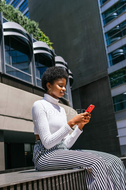 Baixo ângulo de jovem mulher negra em roupas elegantes sentado no banco e celular de navegação no dia ensolarado na rua moderna da cidade — Fotografia de Stock
