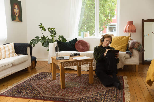 Reflexiva presentadora de radio femenina escribiendo en un cuaderno mientras está sentada en el suelo en casa y grabando podcast - foto de stock