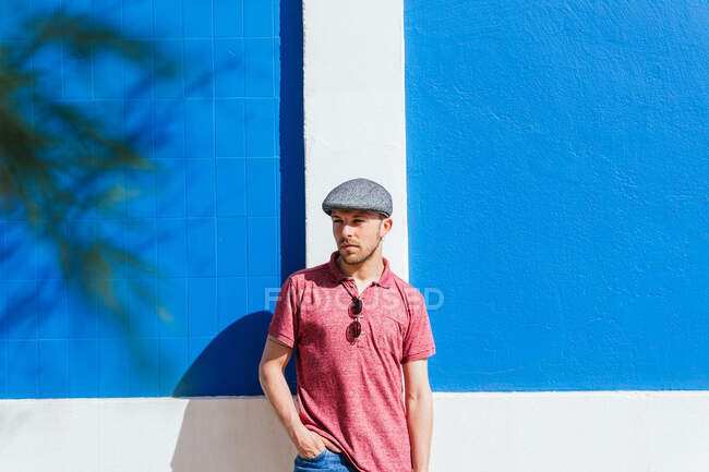 Ragionevole giovane uomo barbuto in elegante abbigliamento casual e cap godendo di giorno d'estate vicino al muro blu sulla strada urbana — Foto stock