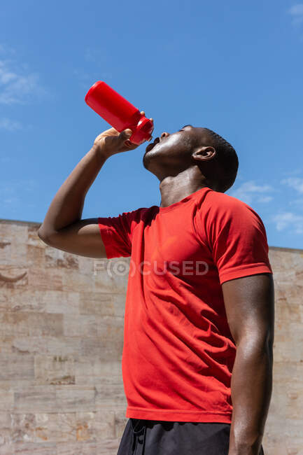 Baixo ângulo de homem atlético afro-americano sedento bebendo água doce de garrafa de plástico durante o treinamento no dia ensolarado na cidade — Fotografia de Stock