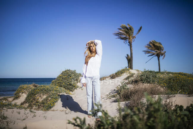 Schöne blonde junge Frau steht an einem sonnigen Tag am Strand in urbaner Kleidung — Stockfoto