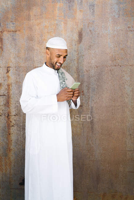 Веселий мусульманин у традиційному одязі посміхається і переглядає мобільний, стоячи біля обшарпаної стіни на вулиці. — стокове фото
