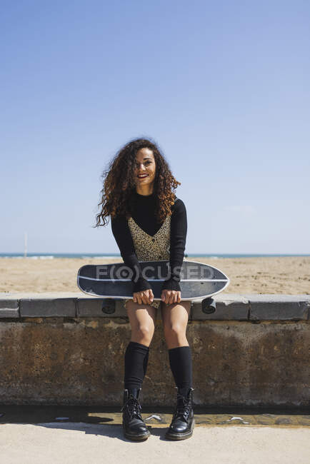 Full body of cheerful female with skateboard in hands looking at camera while sitting on stone border against sandy coast — Stock Photo