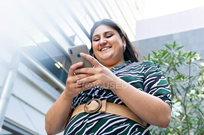 From below of cheerful young curvy female in stylish dress messaging on mobile phone near modern urban building — Stock Photo