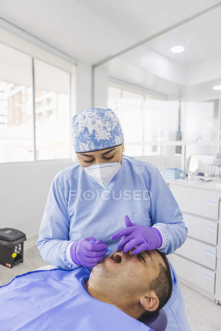 Estomatóloga femenina en uniforme y máscara respiratoria curando dientes de paciente masculino en el hospital - foto de stock