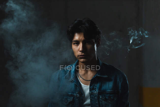 Retrato de jovem latino sério homem olhando com confiança para a câmera em meio a fumaça sob iluminação dramática — Fotografia de Stock